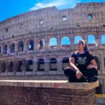 O que fazer em Roma: O céu está azul e o Coliseu está de fundo. Estou sentada em um dos muros que cercam o monumento.