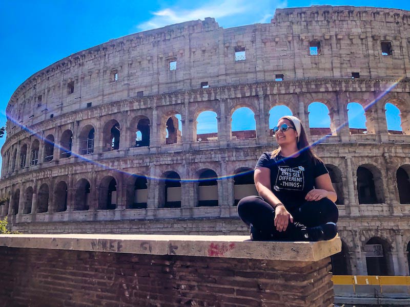 O que fazer em Roma: O céu está azul e o Coliseu está de fundo. Estou sentada em um dos muros que cercam o monumento.